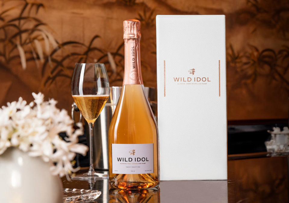 A glass of WILD IDOL Alcohol Free Sparkling Rosé Wine standing on a table next to its bottle and Luxury Gift Box