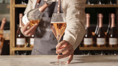 A person in an apron serves a glass of non alcoholic sparkling wine at a bar, with bottles in the background.
