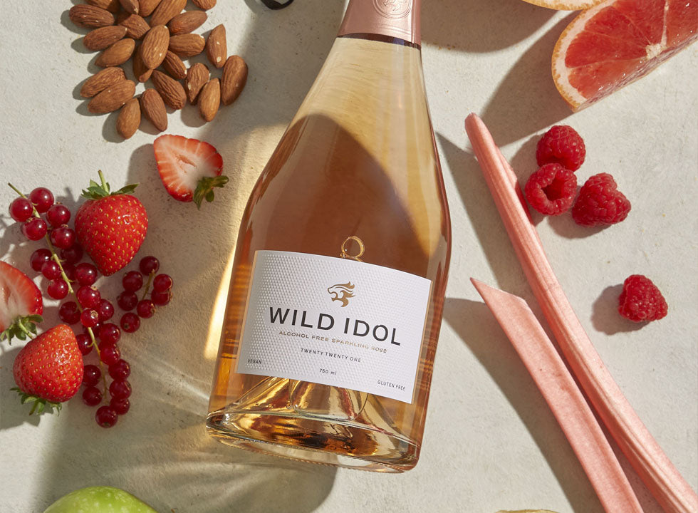 A bottle of Wild Idol Alcohol Free Sparkling Rosé Wine lying next to the fruit that comprise its flavour