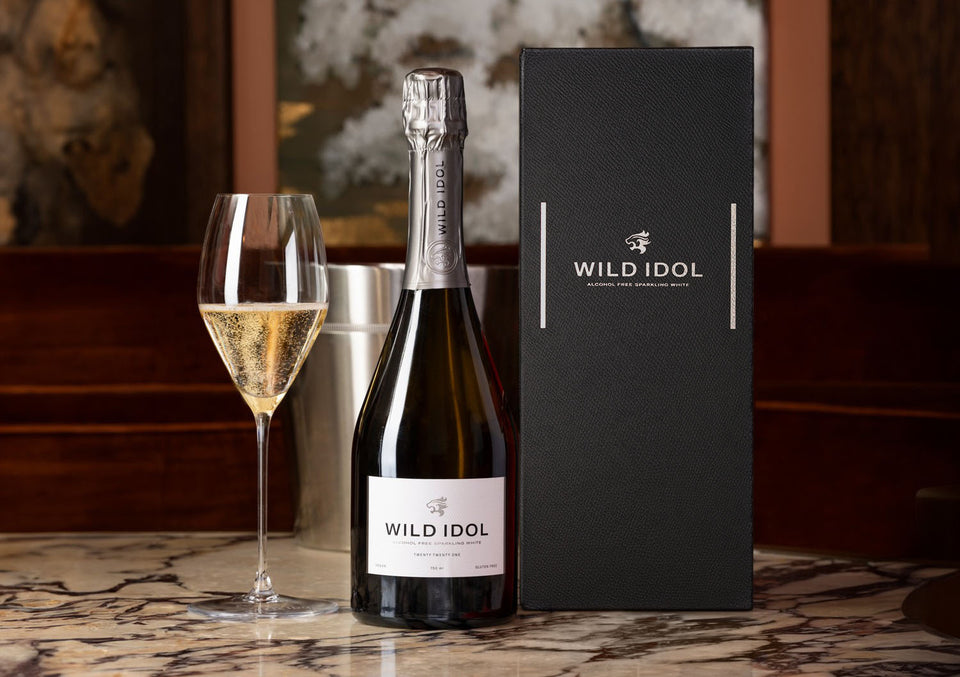A glass of WILD IDOL Alcohol Free Sparkling White Wine standing on a table next to its bottle and Luxury Gift Box
