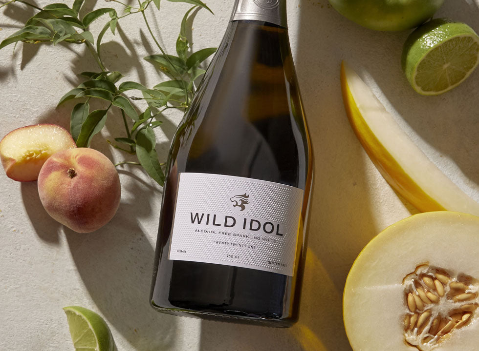 A bottle of Wild Idol Alcohol Free Sparkling White Wine lying next to the fruit that comprise its flavour